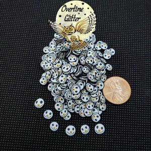 May include: A large pile of white and black smiley face confetti with a yellow dot in the middle. The confetti is scattered on a black surface. A penny is included for scale.