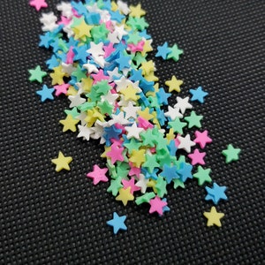 May include: A scattering of colorful star-shaped confetti in shades of pink, yellow, blue, green, and white. The confetti is scattered on a black textured surface, creating a vibrant and playful visual.