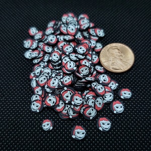 May include: A pile of black, red, and white polymer clay slices shaped like a skull wearing a Santa hat. A penny is included for size reference. The skull slices are scattered on a black surface.