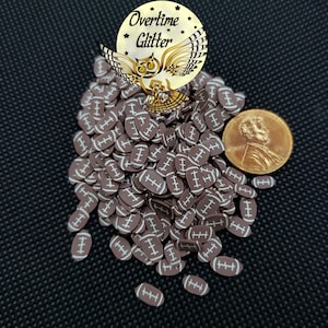 May include: A collection of brown football-shaped polymer clay pieces with white stripes. A penny and a gold-colored owl logo with the text "Overtime Glitter" are also visible. The items are on a black textured surface.