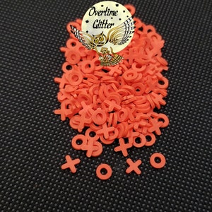May include: A pile of orange X and O shaped craft supplies. The image includes a circular logo with an owl and the words "Overtime Glitter". These small, flat shapes are likely used for crafting or embellishment projects.