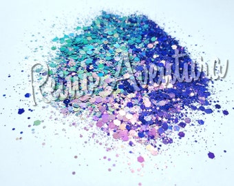 May include: A pile of loose craft glitter featuring a gradient of colors including deep royal blue, bright turquoise, and soft lavender. The glitter consists of various hexagonal and circular shapes of different sizes. The text Reino Aventura is visible.