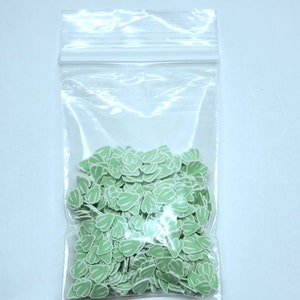 May include: A clear plastic bag filled with small, light green leaf-shaped embellishments with white stripes. The leaves are densely packed inside the bag, creating a textured appearance. The background is a plain white surface.