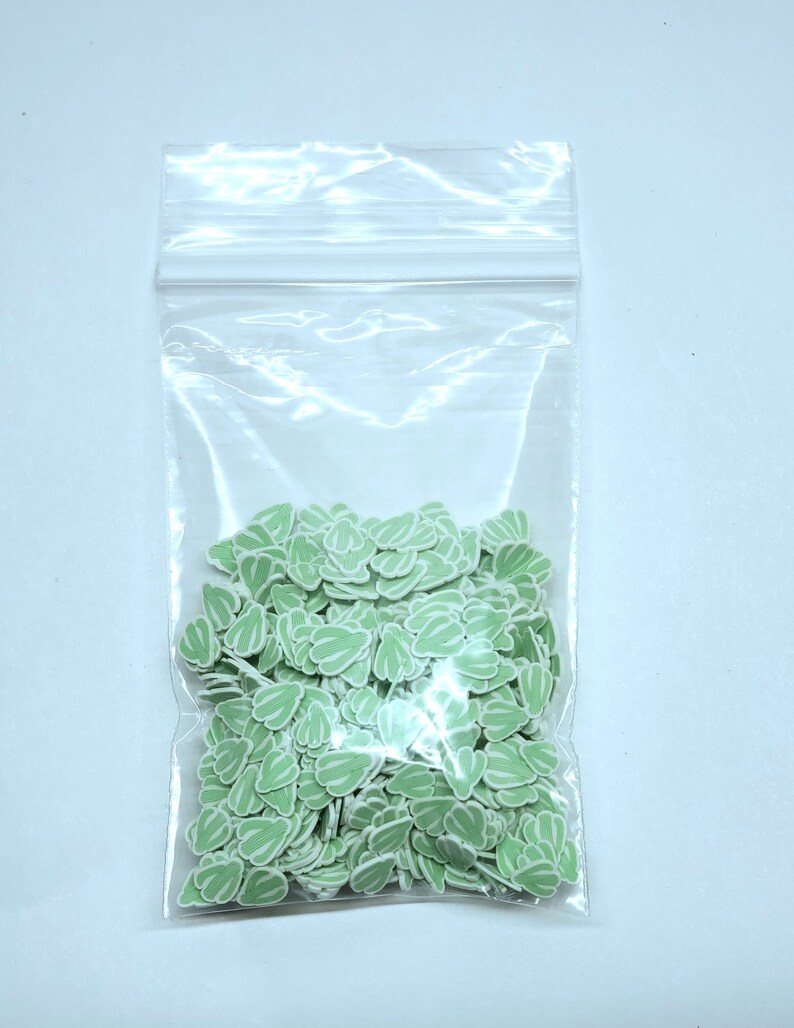 May include: A clear plastic bag filled with small, light green leaf-shaped embellishments with white stripes. The leaves are densely packed inside the bag, creating a textured appearance. The background is a plain white surface.