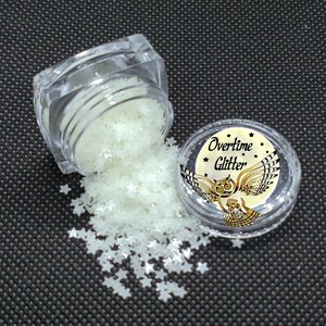 May include: Clear plastic jar spilling star-shaped glitter. The glitter is a pale, glowing green color. A second jar with the text "Overtime Glitter" and an owl graphic is also visible.