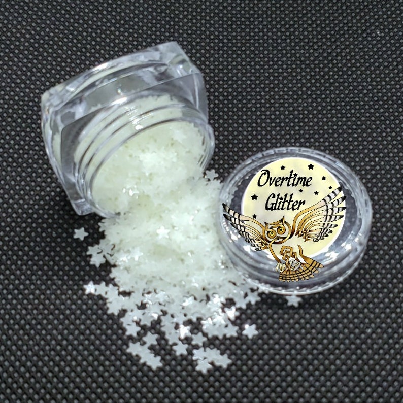 May include: Clear plastic jar spilling star-shaped glitter. The glitter is a pale, glowing green color. A second jar with the text "Overtime Glitter" and an owl graphic is also visible.