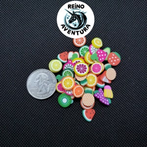 May include: A collection of colorful fruit-shaped polymer clay charms, including watermelon, lemon, orange, kiwi, and strawberry. A US quarter coin is next to the charms for size comparison. The image also includes a logo that says "REINO AVENTURA".