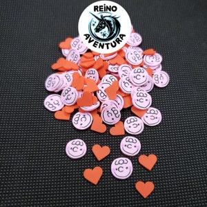 May include: A pile of pink and orange foam shapes. The shapes are hearts and smiley faces. The smiley faces have black eyes and a black mouth. The text "Reino Aventura" is on a white circle with a unicorn and stars.