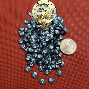 May include: A pile of black skull and crossbones confetti with white details. A gold-colored sticker with an owl and the words "Overtime Glitter" is at the top. A US penny is next to the confetti on a red surface.