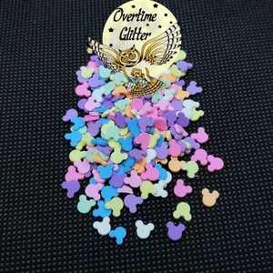 May include: A collection of colorful Mickey Mouse-shaped confetti in pastel shades of pink, purple, blue, yellow, and orange. The confetti is scattered on a black surface, with a gold-colored emblem that reads "Overtime Glitter" and features an owl.