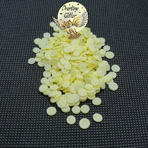 May include: A pile of yellow lemon slice-shaped polymer clay pieces with white details. The pieces are scattered on a dark gray textured surface. A circular logo with an owl and the text "Overtime Glitter" is in the upper left.