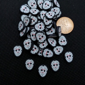 May include: A large pile of white and black Halloween themed charms with a red hockey mask design. The charms are scattered on a black surface. A US penny is included for scale.