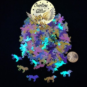 May include: A collection of iridescent unicorn-shaped glitter in shades of purple, blue, and yellow. The glitter is scattered on a black surface, with a penny for size comparison. A gold-colored logo with an owl and the words "Overtime Glitter" is at the top.