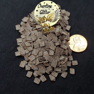 May include: A pile of brown and white square-shaped resin embellishments with a white and green eye design. The embellishments are scattered on a black surface. A US penny is included for scale.