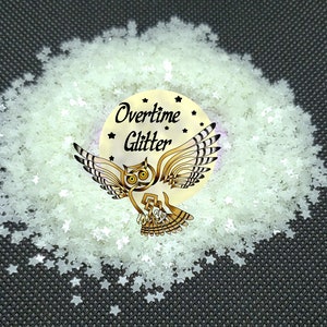 May include: A pile of star-shaped glitter in a pale green color. The glitter surrounds a circular sticker with an owl illustration and the words "Overtime Glitter". The background is a dark gray.