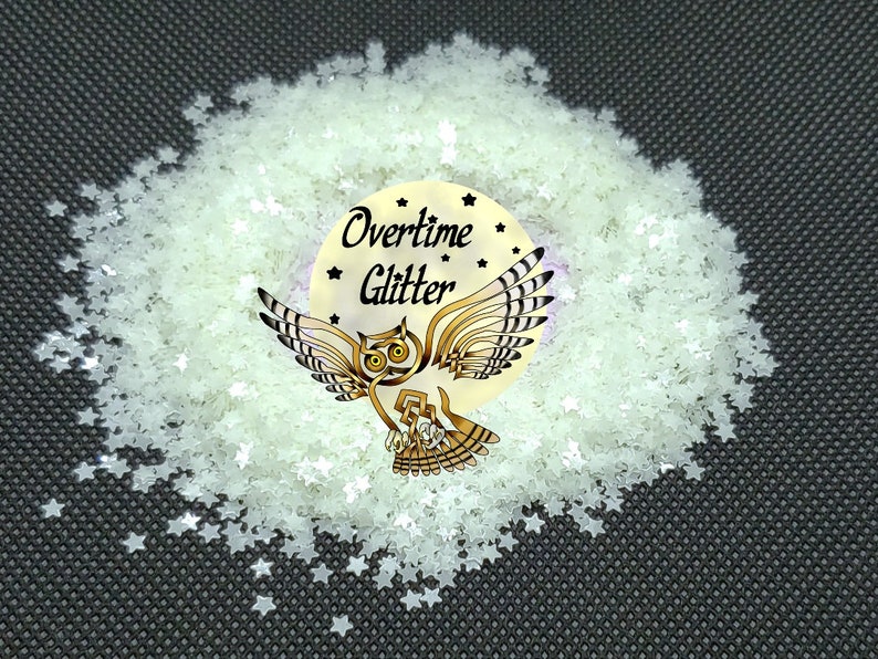 May include: A pile of star-shaped glitter in a pale green color. The glitter surrounds a circular sticker with an owl illustration and the words "Overtime Glitter". The background is a dark gray.