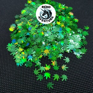May include: Green iridescent glitter shaped like marijuana leaves. The glitter is scattered on a black surface. The image includes a logo that says "Reino Aventura".