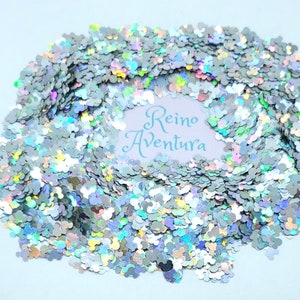 May include: Silver glitter in the shape of a mouse head, scattered across a light blue surface. The glitter has a holographic effect, reflecting multiple colors. The text "Reino Aventura" is visible in the center.