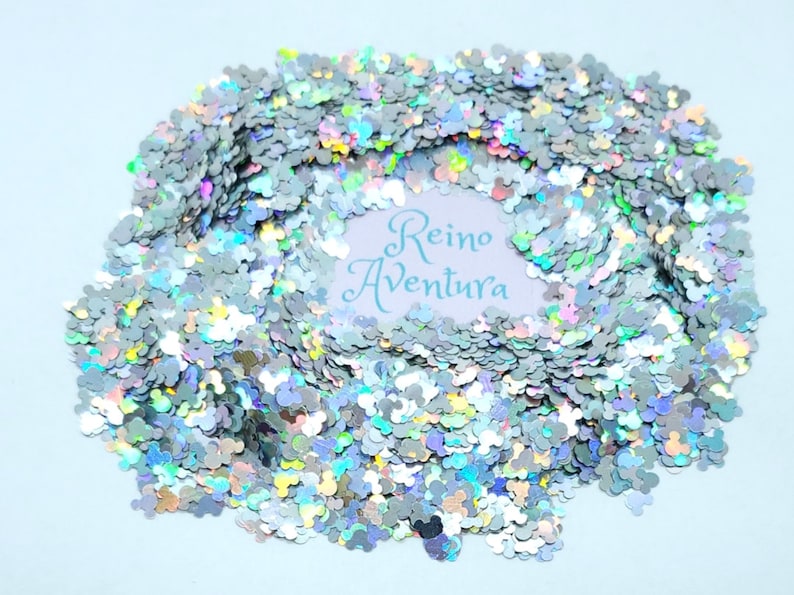 May include: Silver glitter in the shape of a mouse head, scattered across a light blue surface. The glitter has a holographic effect, reflecting multiple colors. The text "Reino Aventura" is visible in the center.