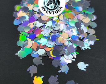 May include: Iridescent glitter in the shape of a hand giving the middle finger. The glitter has a holographic effect, reflecting colors like silver, blue, green, and gold. A circular logo with a unicorn and the text "REINO AVENTURA" is visible.