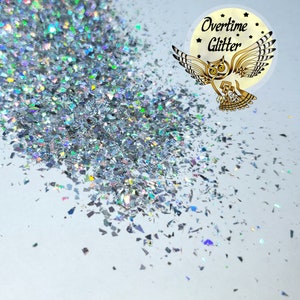 May include: Silver glitter flakes scattered on a white surface. The glitter has a reflective, iridescent quality, catching light to create a shimmering effect. The image includes a logo with an owl and the words "Overtime Glitter".