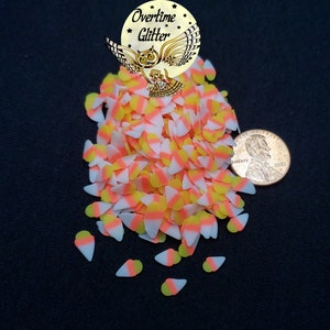 May include: A pile of small, candy corn-shaped sprinkles in white, yellow, and orange. The sprinkles are scattered around a US penny for scale.