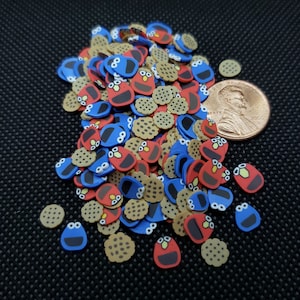 May include: A pile of colorful cookie-shaped resin charms featuring Cookie Monster and Elmo faces, along with brown cookie charms. The charms are scattered on a black surface.