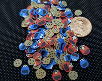 Pode incluir: Um monte de pingentes de resina em forma de biscoito com rostos de Cookie Monster e Elmo, juntamente com pingentes de biscoito marrons. Os pingentes estão espalhados sobre uma superfície preta.