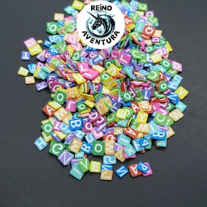 May include: A pile of colorful alphabet letter tiles in various colors, including pink, green, blue, and yellow. Each tile has a different letter of the alphabet. The image also includes a circular logo with a unicorn and the text "REINO AVENTURA".