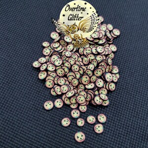 May include: A large pile of small, round, yellow and orange pumpkin-shaped resin embellishments with black outlines. The embellishments are scattered on a dark gray textured surface. The text "Overtime Glitter" is visible in the top left corner of the image.
