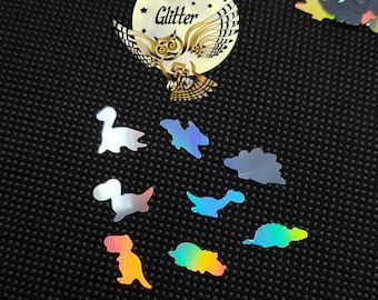 May include: A collection of iridescent dinosaur-shaped glitter pieces on a black textured surface. The glitter pieces are in various colors, including silver, blue, and yellow. A gold-colored sticker with an owl and the words "Overtime Glitter" is also present.