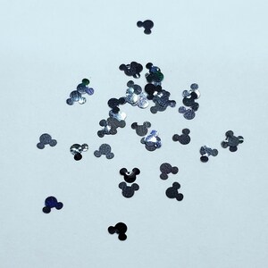 Pode incluir: Confetti de glitter prateado em forma de cabe&ccedil;as do Mickey Mouse espalhados sobre um fundo branco.