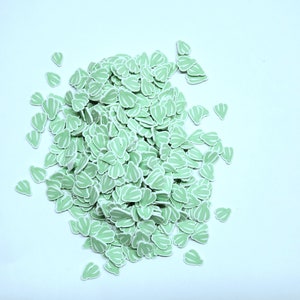 May include: A pile of light green, leaf-shaped embellishments with white striped details. These small, decorative pieces are scattered on a white surface. The leaves have a rounded shape and are likely used for crafting or scrapbooking.