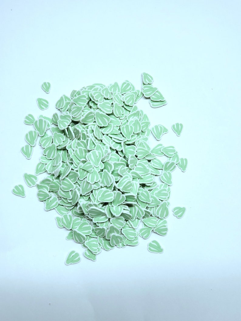 May include: A pile of light green, leaf-shaped embellishments with white striped details. These small, decorative pieces are scattered on a white surface. The leaves have a rounded shape and are likely used for crafting or scrapbooking.