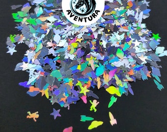 May include: Iridescent glitter in various shapes, including stars, rockets, and figures, scattered on a black surface. The glitter reflects multiple colors, creating a shimmering effect. The image includes a circular logo with a unicorn and the text "REINO AVENTURA".