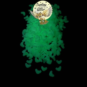 May include: Green glow-in-the-dark glitter in the shape of bats. The glitter is scattered on a black background. The image also includes a logo with an owl and the text "Overtime Glitter".