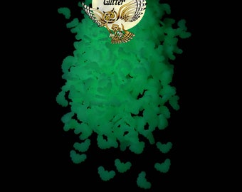May include: Green glow-in-the-dark glitter in the shape of bats. The glitter is scattered on a black background. The image also includes a logo with an owl and the text "Overtime Glitter".