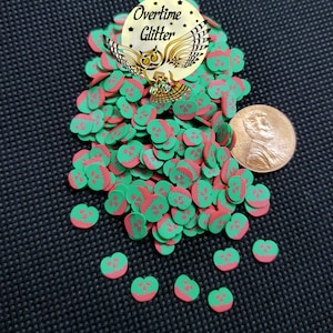 May include: A large pile of green and red apple-shaped sprinkles with a white outline and a small bite mark. The sprinkles are scattered on a black surface. A penny is included for scale.
