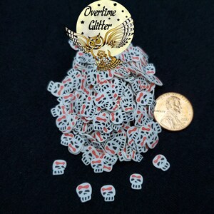 May include: A large pile of white and red skull-shaped sprinkles with a red crossbones design. The sprinkles are next to a US penny for size comparison.