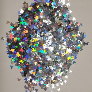 May include: A pile of silver glitter shaped like Mickey Mouse heads. The glitter has a holographic effect, reflecting multiple colors. The glitter is scattered on a light gray surface.