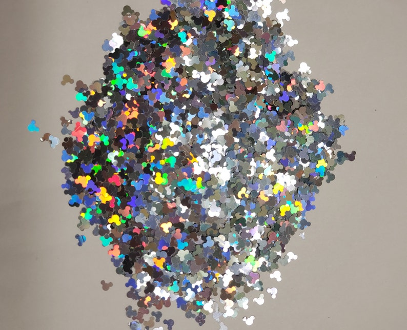May include: A pile of silver glitter shaped like Mickey Mouse heads. The glitter has a holographic effect, reflecting multiple colors. The glitter is scattered on a light gray surface.