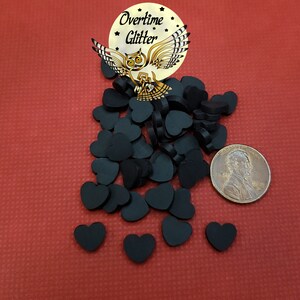 May include: A pile of black wooden heart-shaped embellishments, approximately the size of a US penny, on a red background. The image also includes a small sticker with an owl and the text "Overtime Glitter".