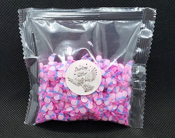 Pode incluir: Um saco plástico transparente cheio de granulado de argila polimérica rosa, branca e azul. Um adesivo branco com o texto "Overtime Glitter" e um gráfico de coruja está no saco.