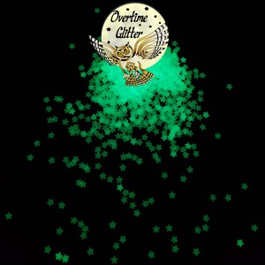 May include: Black background with scattered green star-shaped glitter. A circular logo features an owl with outstretched wings, the text "Overtime Glitter" and a crescent moon. The glitter appears to glow.