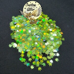 May include: A pile of green glitter shaped like cannabis leaves. The glitter has a holographic effect, reflecting multiple colors. A circular sticker with an owl graphic and the text "Overtime Glitter" is in the upper portion of the image.