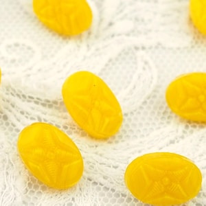 May include: A collection of bright yellow, oval-shaped beads with a textured floral design. The beads are scattered on a white lace fabric, creating a visually appealing contrast of colors and textures. These beads could be used for jewelry making or other craft projects.