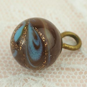 May include: A round button with a brown base, swirled with blue and gold accents. It has a small, brass-coloured loop on the back. The button is set against a soft, off-white lace background.