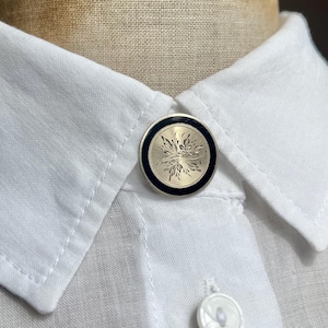 May include: Close-up of a white collared shirt with a decorative button. The button is round with a silver floral design and a black border. A second white button is visible below the decorative button.