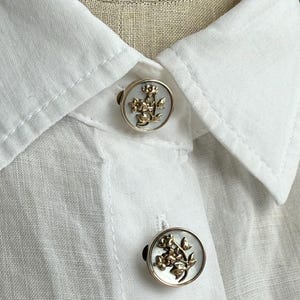 以下が含まれることがあります： 白いシャツのクローズアップ。2つの装飾ボタンが付いています。ボタンは丸く、白い背景に金色の花の模様があり、銀色のフレームにセットされています。シャツは軽くて質感のある生地で作られています。