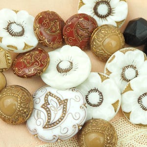 May include: A selection of vintage buttons in diverse shapes and sizes. The buttons display colours such as white, gold, red, and brown. Some buttons have floral designs, while others have intricate patterns and textures. There are also black faceted beads.